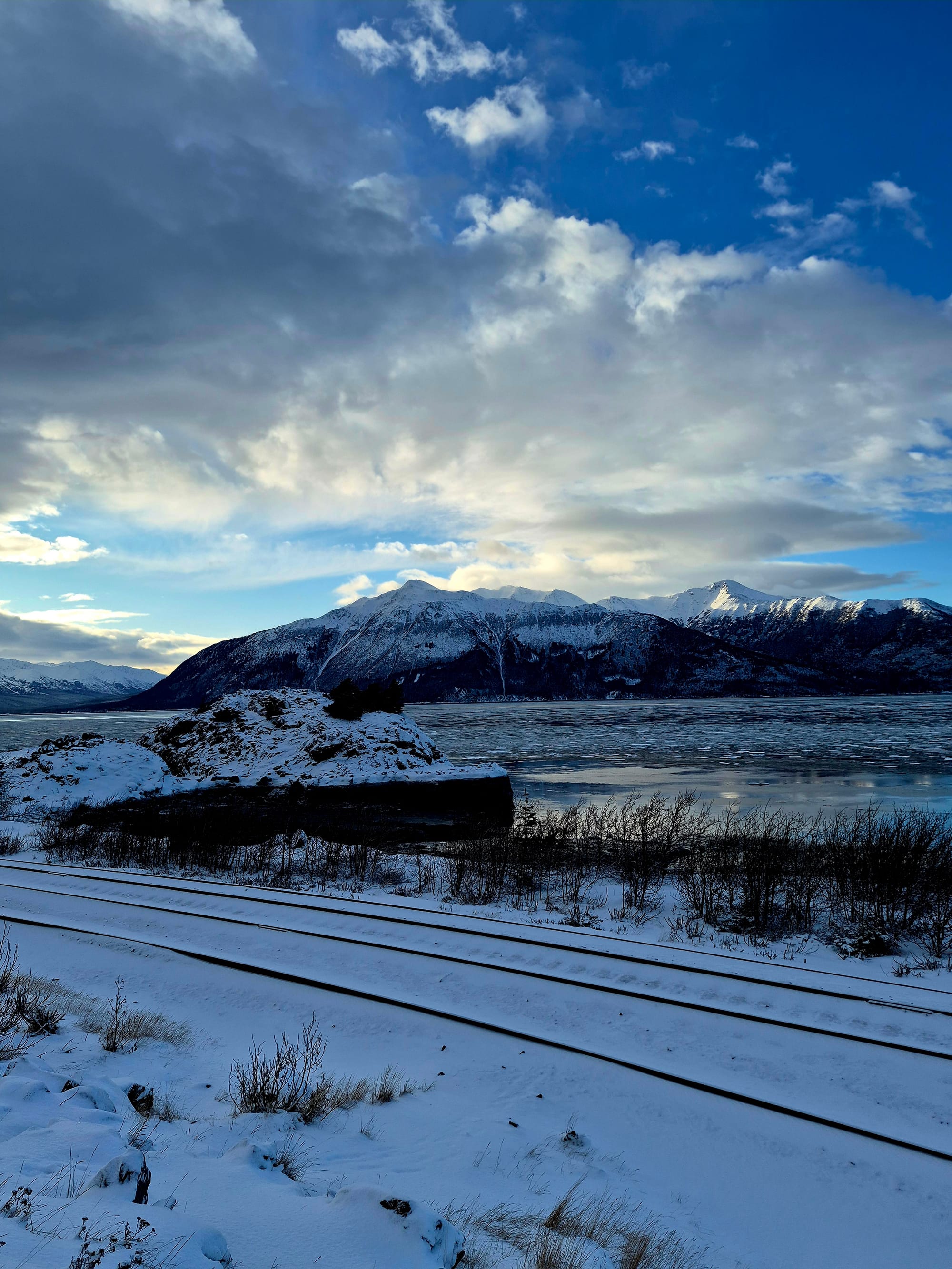 Turnagain Arm in winter — snow-lined rail tracks, tidal flats locked in ice, and the Chugach Mountains rising sharply from the water beneath a restless Alaska sky.